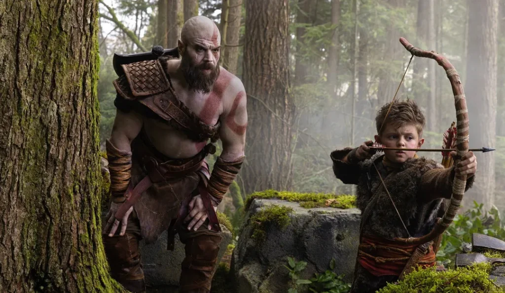 Skallig man med skägg i vikingakläder med yxa står bredvid pojke som håller upp båge i skogsmiljö med mossa och träd.